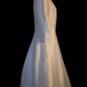 May include: A white wedding dress with long sleeves and a full skirt. The dress features delicate beaded embellishments along the sleeves and bodice. The dress is made of a smooth, flowing fabric.