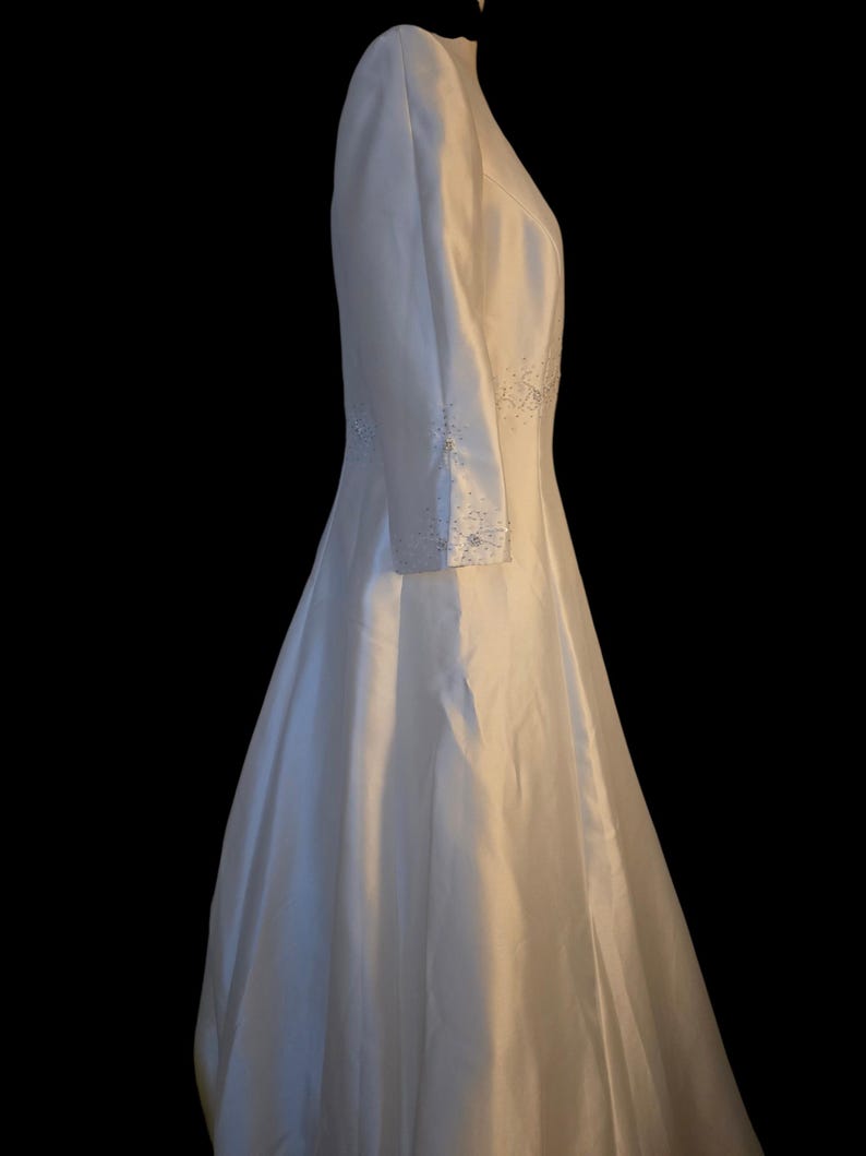 May include: A white wedding dress with long sleeves and a full skirt. The dress features delicate beaded embellishments along the sleeves and bodice. The dress is made of a smooth, flowing fabric.