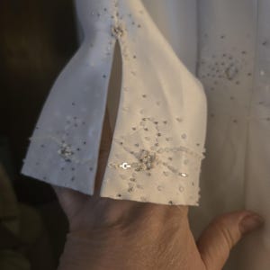 May include: Close-up of a white garment sleeve with a split cuff. The cuff is embellished with small, scattered silver beads and a cluster of larger, iridescent sequins. A small, decorative button is at the cuff's center.