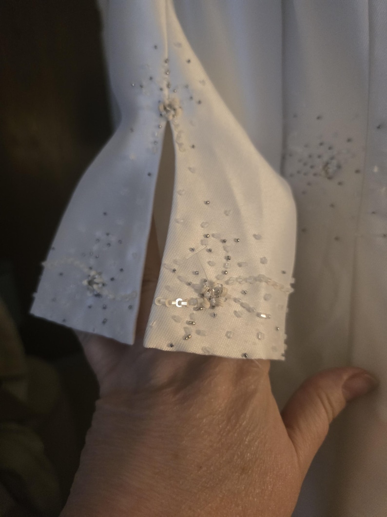 May include: Close-up of a white garment sleeve with a split cuff. The cuff is embellished with small, scattered silver beads and a cluster of larger, iridescent sequins. A small, decorative button is at the cuff's center.