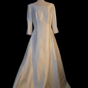 May include: A white wedding dress with a boat neckline and three-quarter sleeves. The dress features a fitted bodice with a subtle beaded embellishment at the waist and a flowing A-line skirt. The dress is displayed on a mannequin against a black background.