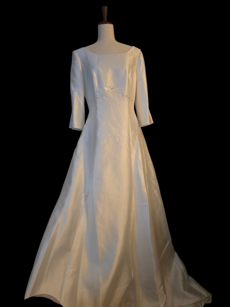 May include: A white wedding dress with a boat neckline and three-quarter sleeves. The dress features a fitted bodice with a subtle beaded embellishment at the waist and a flowing A-line skirt. The dress is displayed on a mannequin against a black background.