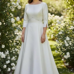 May include: A white wedding dress with a boat neckline and three-quarter sleeves. The dress features a detailed waistline and a flowing A-line skirt. The bride is standing in a garden with white roses.
