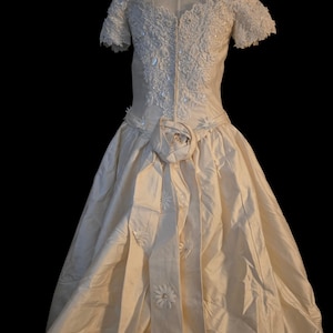 May include: An off-white, floor-length gown featuring an off-the-shoulder neckline with short sleeves. The bodice is adorned with intricate floral lace and beadwork. A large fabric rose sits at the waist, with a long sash featuring floral accents.