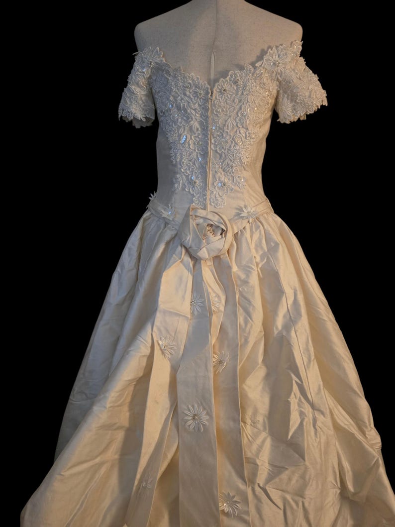 May include: An off-white, floor-length gown featuring an off-the-shoulder neckline with short sleeves. The bodice is adorned with intricate floral lace and beadwork. A large fabric rose sits at the waist, with a long sash featuring floral accents.