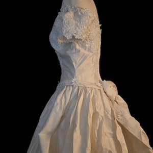 May include: An off-white, floor-length gown featuring an off-the-shoulder neckline with intricate white floral lace and pearl embellishments. The bodice transitions into a full, gathered skirt adorned with small fabric daisies along the waistline.