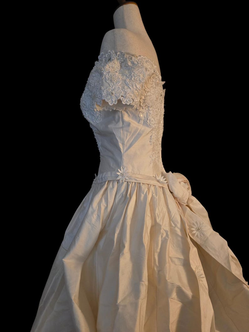 May include: An off-white, floor-length gown featuring an off-the-shoulder neckline with intricate white floral lace and pearl embellishments. The bodice transitions into a full, gathered skirt adorned with small fabric daisies along the waistline.