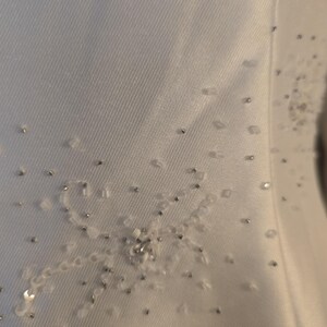 May include: Close-up of a white fabric with delicate embellishments. The design features a floral pattern created with small, clear beads and silver accents. The fabric has a subtle texture, adding depth to the design.