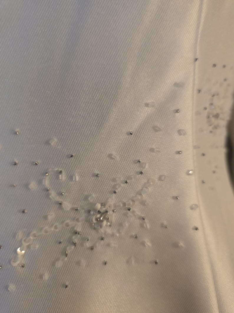 May include: Close-up of a white fabric with delicate embellishments. The design features a floral pattern created with small, clear beads and silver accents. The fabric has a subtle texture, adding depth to the design.