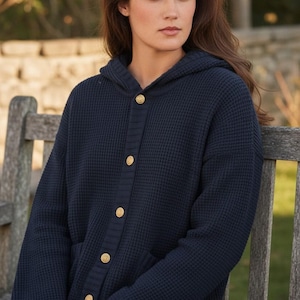 May include: A navy blue hooded cardigan featuring a waffle knit texture, a front button closure with gold-toned metallic buttons, and long sleeves with small gold-toned zippers at the cuffs. The garment has a ribbed placket and ribbed sleeve cuffs.
