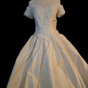 May include: An off-the-shoulder ivory gown featuring a sweetheart neckline and short sleeves. The bodice is adorned with intricate white lace, floral embroidery, and sequins. A V-shaped waistline is accented with small floral appliqu&eacute;s.