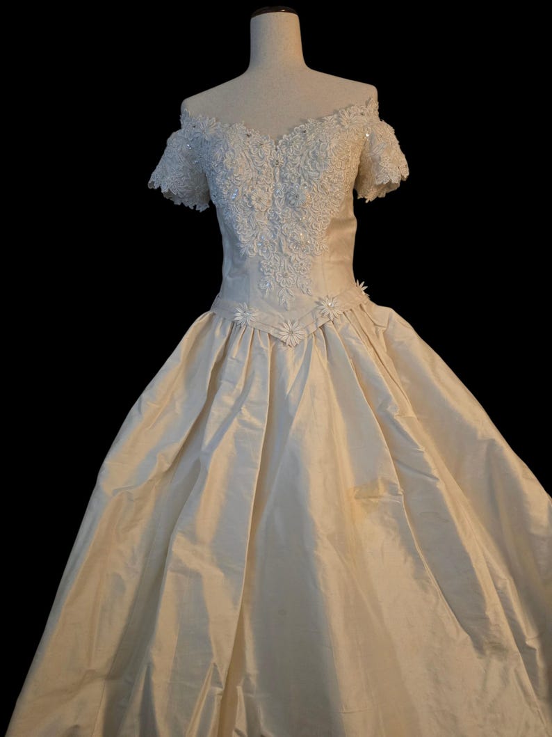 May include: An off-the-shoulder ivory gown featuring a sweetheart neckline and short sleeves. The bodice is adorned with intricate white lace, floral embroidery, and sequins. A V-shaped waistline is accented with small floral appliqu&eacute;s.