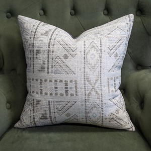 May include: A square throw pillow featuring a textured white fabric with a woven pattern of gray and light blue geometric shapes, including vertical stripes, diamonds, and chevron lines arranged in a repeating, symmetrical grid-like layout.