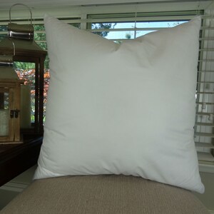 Puede incluir: Una almohada blanca cuadrada grande con una apariencia suave y esponjosa. La almohada est&aacute; hecha de una tela lisa y est&aacute; colocada sobre una superficie de tono neutro, probablemente un sof&aacute; o una silla. La almohada es el foco principal de la imagen.