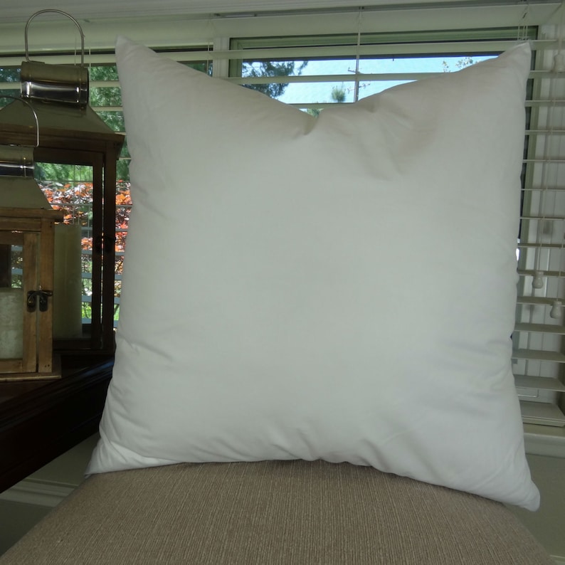 Puede incluir: Una almohada blanca cuadrada grande con una apariencia suave y esponjosa. La almohada est&aacute; hecha de una tela lisa y est&aacute; colocada sobre una superficie de tono neutro, probablemente un sof&aacute; o una silla. La almohada es el foco principal de la imagen.