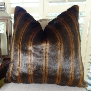 May include: A brown and gold striped faux fur throw pillow. The pillow is made of soft, plush fabric and has a luxurious look and feel.