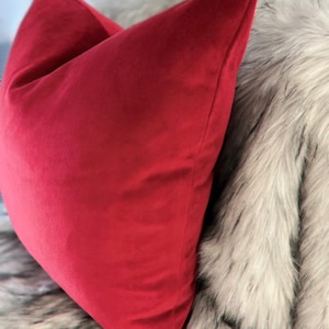 May include: A red velvet pillow on a white faux fur throw. The pillow is a solid color and has a simple, rectangular shape.