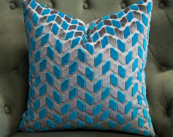 May include: A square throw pillow featuring a repeating geometric chevron pattern. The design consists of alternating rows of raised, textured bright turquoise blue and soft grey velvet-like fabric segments arranged in a vertical zigzag formation.