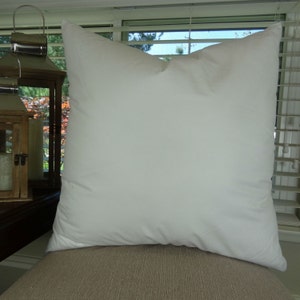 May include: A large, square white pillow with a soft, puffy appearance. The pillow is made of a smooth fabric and is positioned on a neutral-toned surface. The pillow is the primary focus of the image.