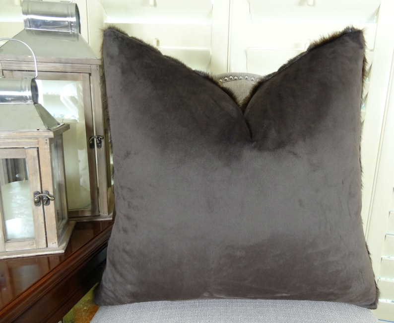 May include: A brown faux fur throw pillow with a square shape and a subtle sheen. The pillow has a brown trim around the edges.