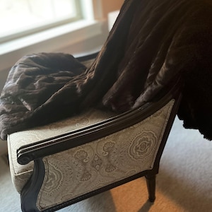May include: A brown faux fur throw draped over a brown wooden armchair with a beige paisley patterned fabric seat and back.