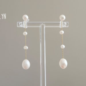May include: A pair of pearl drop earrings with a gold chain and a pearl at the bottom. The earrings are on a display stand.