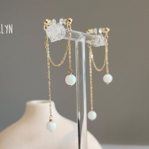 May include: A pair of gold earrings with a delicate chain and a white opal gemstone dangling from each earring.