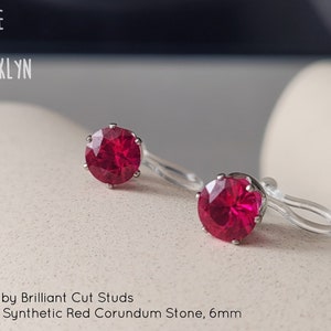 May include: A pair of red ruby brilliant cut stud earrings. The earrings feature a 6mm AAAAA synthetic red corundum stone. The brand name "ALICE IN BROOKLYN" is in the upper left corner.