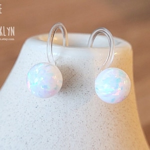 May include: A pair of white opal stud earrings with clear plastic hooks. The earrings are round and have a pearlescent, iridescent sheen. The earrings are displayed on a white ceramic surface. The text "ALICE IN BROOKLYN" and a website address are visible.