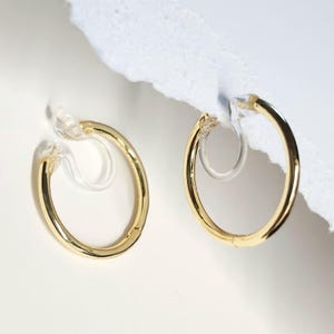 May include: A pair of gold-toned hoop earrings featuring a smooth, circular metal design. Each earring is equipped with a clear, flexible plastic clip-on mechanism attached to the inner curve, allowing for secure placement without a pierced earlobe.