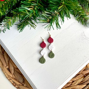May include: A pair of dangle earrings with three round ornaments. The top ornament is red, the middle is white with a snowflake design, and the bottom is green with a forest design.
