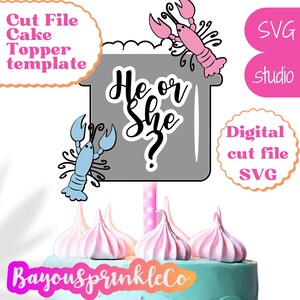 May include: A gray cake topper with the text "He or She?" in black script.  The topper features a pink and blue lobster on either side.  The topper is on top of a cake with pink frosting and white meringue.