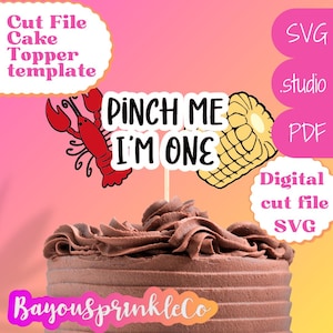 May include: A chocolate cake with a cake topper that says "Pinch Me I'm One" with a red crawfish and a yellow ear of corn on either side. The cake topper is for a first birthday party.