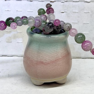 May include: A small, pastel-colored ceramic pot with a crackle glaze, holding a strand of colorful, round beads. The beads are a mix of pink, green, and clear, and are strung on a wire. The pot has a pink and green gradient.