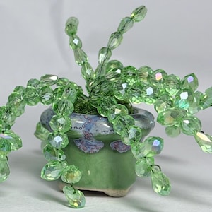 May include: A handcrafted, beaded succulent plant in a small, green ceramic pot. The plant features light green, faceted beads arranged to resemble leaves and stems. The pot has a blue and white speckled design.
