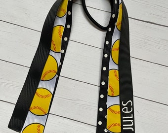 May include: A black and white ribbon with a softball pattern and black polka dots. The ribbon is tied with a black elastic hair tie. The ribbon has the name "Jules" written on it.