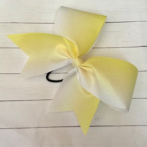 May include: A large yellow and white ombre cheer bow with glitter. The bow is tied with a black elastic band.