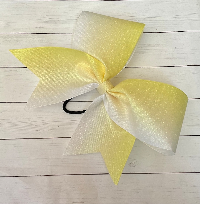May include: A large yellow and white ombre cheer bow with glitter. The bow is tied with a black elastic band.