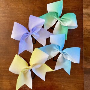 May include: Four ombre cheer bows in different colors. The bows are made of a sparkly fabric and are tied with black elastic. The bows are arranged in a semi-circle on a wooden surface.