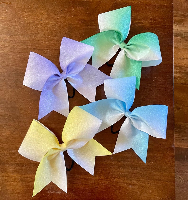 May include: Four ombre cheer bows in different colors. The bows are made of a sparkly fabric and are tied with black elastic. The bows are arranged in a semi-circle on a wooden surface.