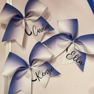 Może przedstawiać: Trzy białe i niebieskie ombre wstążki cheerleaderskie z czarnym napisem. Wstążki są spersonalizowane z nazwami "Caroline", "Ella" i "Kenzie".