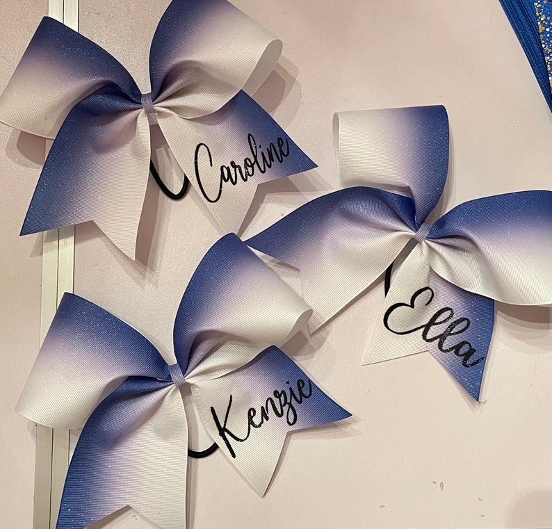 Może przedstawiać: Trzy białe i niebieskie ombre wstążki cheerleaderskie z czarnym napisem. Wstążki są spersonalizowane z nazwami "Caroline", "Ella" i "Kenzie".