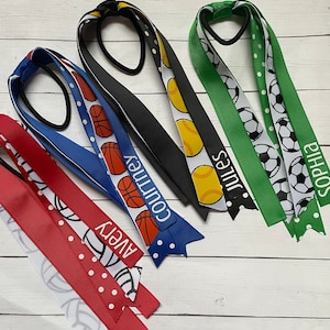 May include: Four colourful hair ties with different sports ball designs and names printed on them. The hair ties are red, blue, black, and green with white polka dots. The names on the hair ties are Avery, Courtney, Jules, and Sophia.