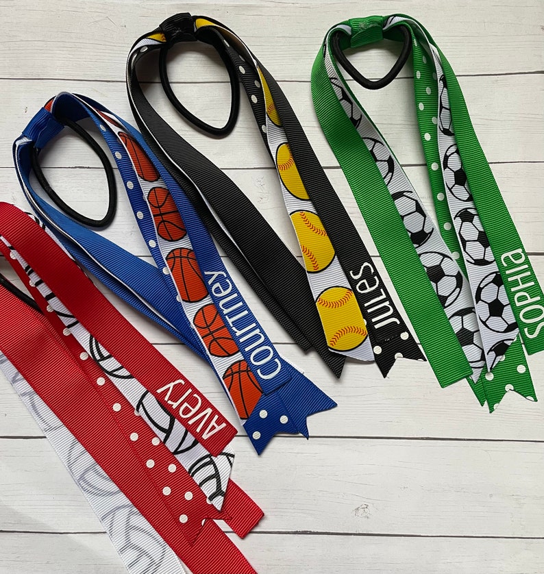 May include: Four colourful hair ties with different sports ball designs and names printed on them. The hair ties are red, blue, black, and green with white polka dots. The names on the hair ties are Avery, Courtney, Jules, and Sophia.