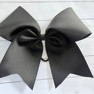 May include: A large black cheer bow with a simple design. The bow is made of a shiny, textured fabric and is tied with a black elastic band.