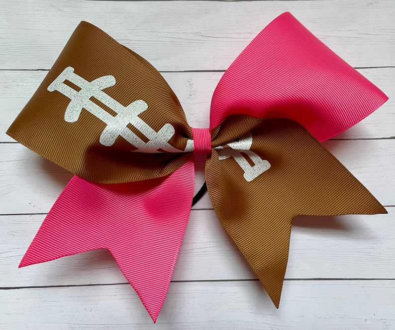 May include: A brown and pink cheer bow with a white football design. The bow is made of two layers of ribbon, with the top layer being brown and the bottom layer being pink.