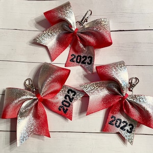 May include: Three red and silver glitter cheer bows with keychains. Each bow has the year 2023 embroidered on it.