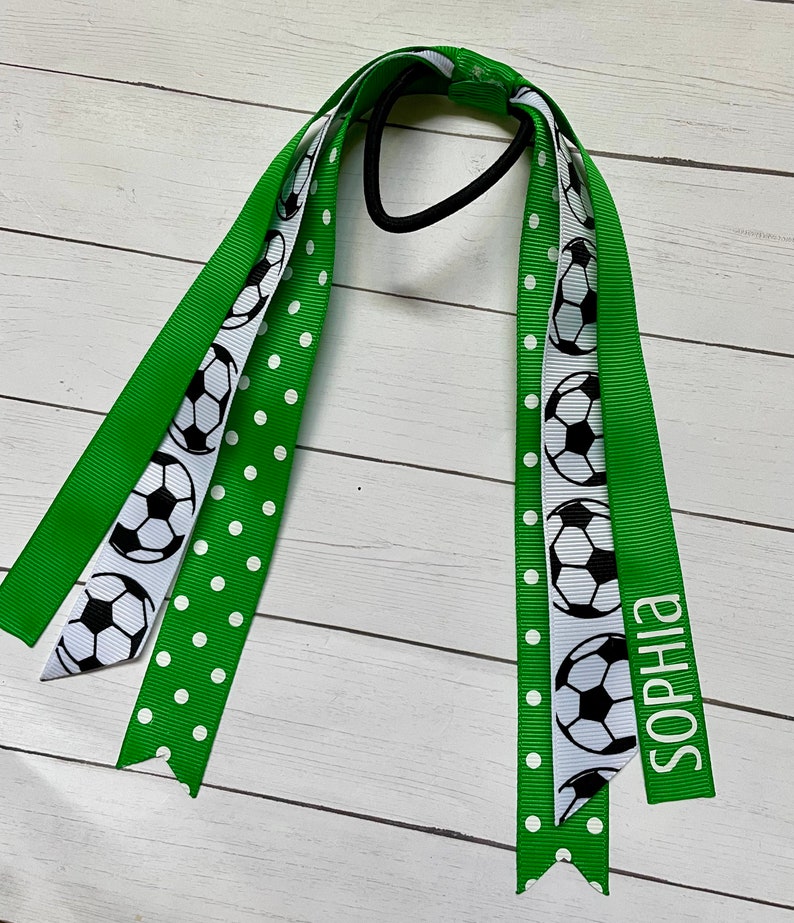 May include: Green and white hair tie with a football pattern and white polka dots. The hair tie has the name "Sophia" written on it.