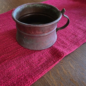 May include: An antique copper mug with a handle, showing signs of age and wear. The mug sits on a textured red runner, placed on a wooden surface. The mug's exterior has a weathered patina, adding to its vintage appeal.