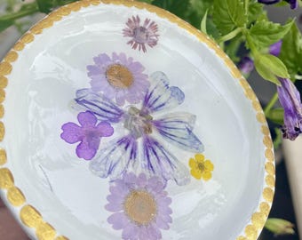 May include: A white, oval-shaped trinket dish with a gold painted rim. The dish is decorated with pressed purple and yellow flowers. The flowers are arranged in the center of the dish, creating a unique and artistic design.